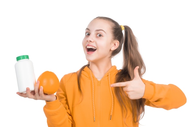 Glückliches Kind zeigt mit dem Finger auf Orangenfrucht und Vitaminpille in einer Glasflasche, isoliert auf weißem Hintergrund. Gesundheit