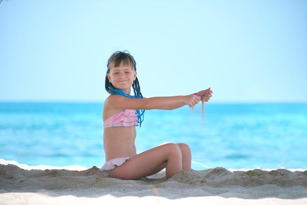 Glückliches Kindermädchen im Bikini-Badeanzug, das während der tropischen Sommerferien mit Sand am Strand spielt
