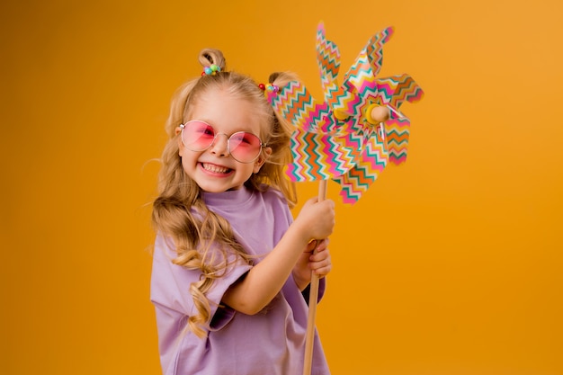 glückliches Kindermädchen in der Sonnenbrille, die eine Babywindmühle auf einem gelben Raum hält