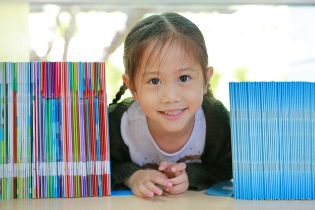 Foto glückliches kleines asiatisches kindermädchen, das auf bücherregal an der bibliothek liegt