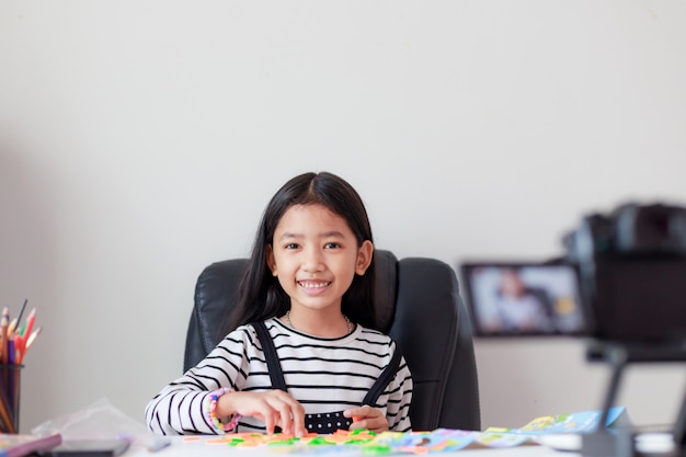 Glückliches kleines asiatisches Mädchen, das am weißen Tisch und am Live-Streaming für Social Media mit Glück sitzt
