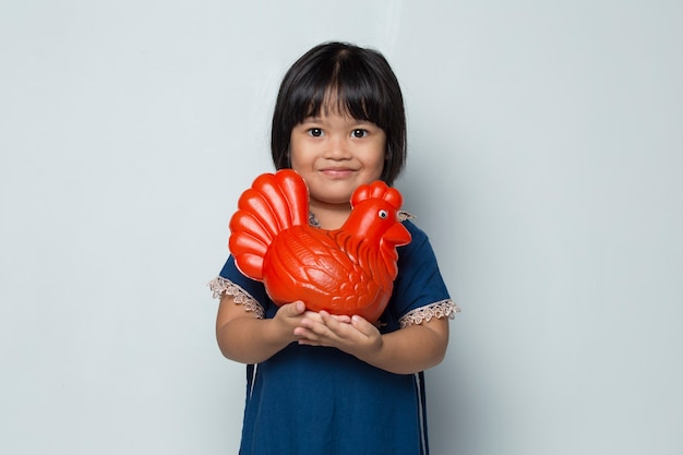 Glückliches kleines asiatisches Mädchen, das Geld mit einer Hühnerbank lokalisiert auf weißem Hintergrund spart