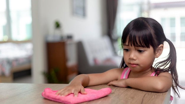 Glückliches kleines asiatisches Mädchen, das lernt, mit einem Lappen im Wohnzimmer zu Hause Hausarbeit und Haushaltskonzept zu reinigen