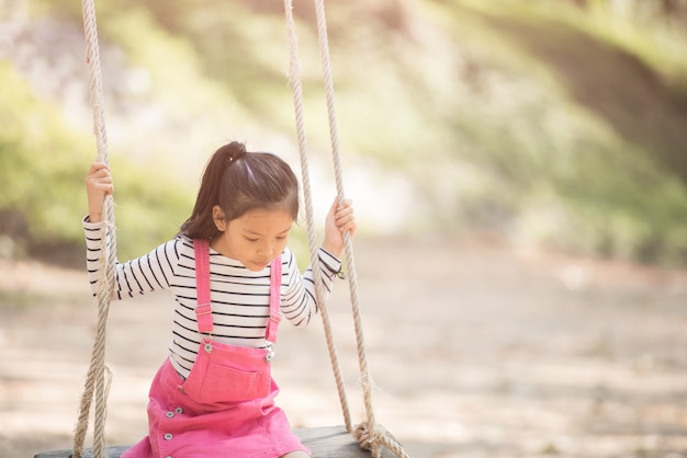 Glückliches kleines asiatisches Mädchenkind, das Spaß hat, auf dem Spielplatz in der Sommerzeit mit Lächeln und gesundem Lachen zu spielen, entzückendes Mädchen, das Spaß auf einer Schaukel im Sommer hat.