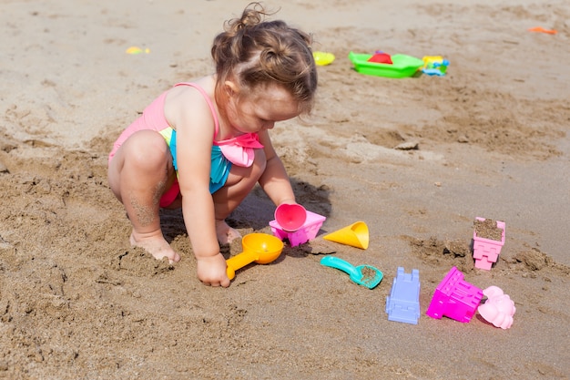 Glückliches kleines Baby im Badeanzug, der im Sand auf dem Strand an einem sonnigen warmen Tag spielt.