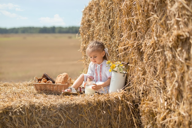 glückliches kleines Baby in Volkskleidung sitzt auf einem Weizenfeld mit einem Glas frischer Milch.