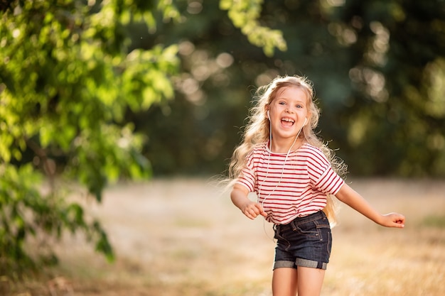 Glückliches kleines blondes Mädchen, das Musik mit Kopfhörern hört, in der Natur im Park tanzt und singt.