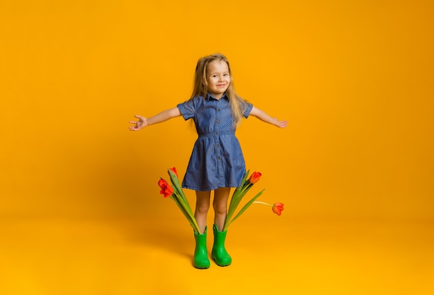 Glückliches kleines blondes Mädchen in einem blauen Kleid und Gummistiefeln steht mit Tulpen auf einer gelben Wand