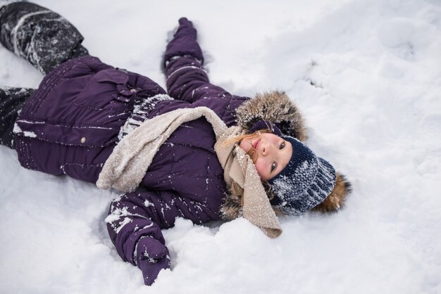 Glückliches kleines blondes Mädchen liegt im Schnee, Kind, das Spaß hat, mit Schnee im Wintertag zu spielen