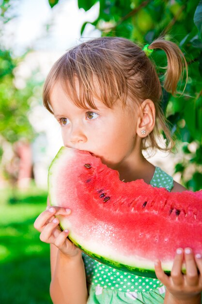 Glückliches kleines blondes Mädchen mit großer Scheibe Wassermelone in der Sommerzeit im Park, im Freien.