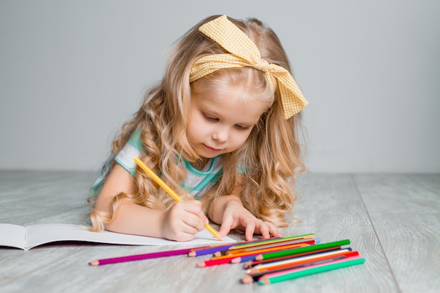 Glückliches kleines Kind, ein charmantes blondes Baby liegt bequem auf dem Holzboden und zeichnet mit Buntstiften auf Papier