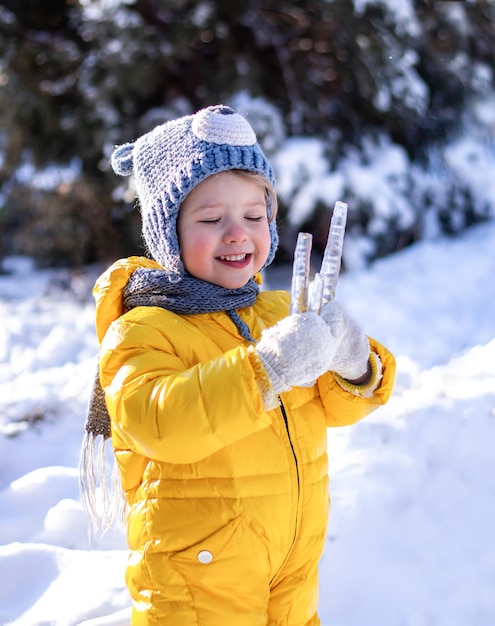 glückliches kleines kind mit geschlossenen augen im gelben schneeanzug, der an sonnigen wintertagen eiszapfen in den händen hält