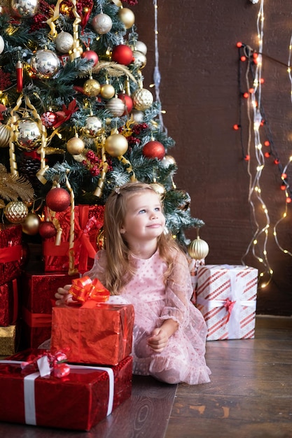 Glückliches kleines lächelndes blondes Mädchen im schönen Kleid mit Geschenkbox nahe Weihnachtsbaum