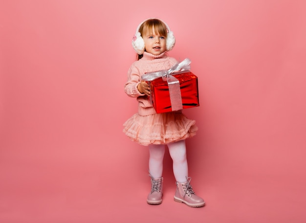 Glückliches kleines lächelndes Mädchen in den Winterkopfhörern mit einer Weihnachtsgeschenkbox.