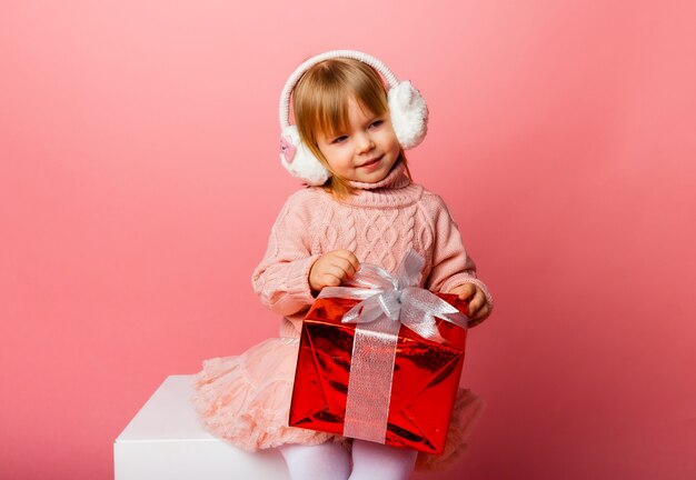 Glückliches kleines lächelndes Mädchen in den Winterkopfhörern mit einer Weihnachtsgeschenkbox.