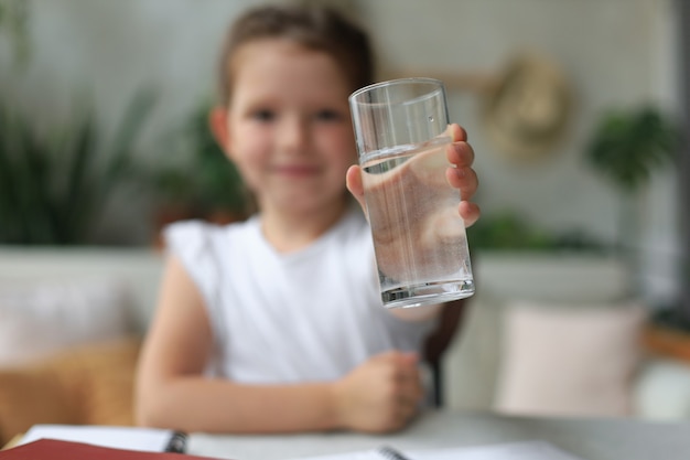 Glückliches kleines Mädchen bietet kristallklares Mineralwasser aus Glas an, kleines Kind empfiehlt die tägliche Dosis sauberen Wassers.