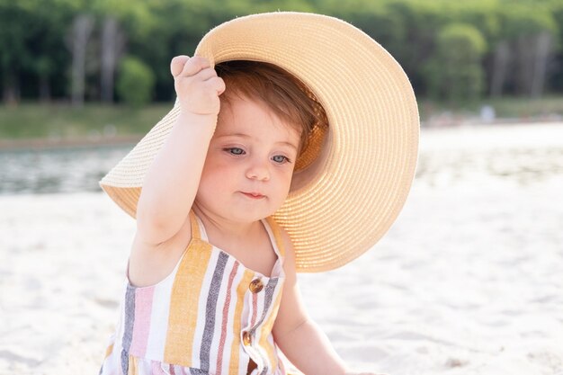 glückliches kleines Mädchen, das an einem sonnigen Tag am Strand auf Sand in gestreiftem Sommerkleid und Strohhut sitzt