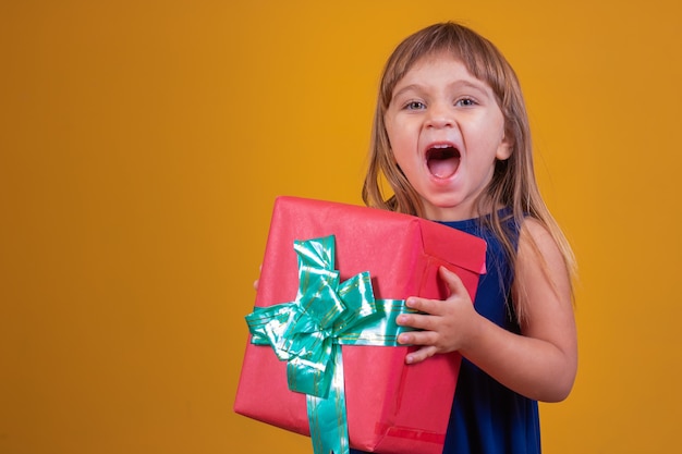 Glückliches kleines Mädchen, das ein Geschenk auf gelbem Hintergrund hält.