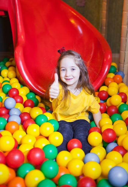 Glückliches kleines Mädchen, das im Kindergarten mit bunten Bällen spielt und Spaß hat und Daumen oben im Spielzentrum zeigt