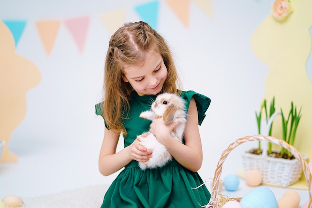 Glückliches kleines Mädchen, das niedlichen flauschigen Hasen nahe gemalten Ostereiern hält