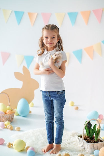 Glückliches kleines Mädchen, das niedlichen flauschigen Hasen nahe gemalten Ostereiern hält