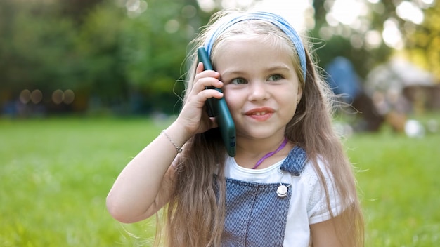 Glückliches kleines Mädchen, das sich im Sommerpark auf ihrem Verkaufstelefon unterhält