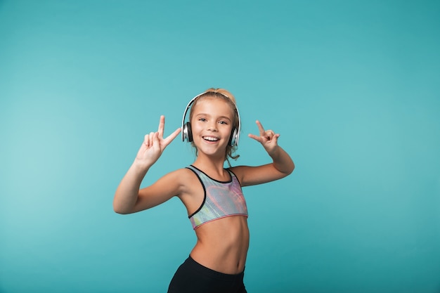 Glückliches kleines Mädchen, das Sportkleidung trägt, die Musik mit Kopfhörern lokalisiert über blauer Wand hört