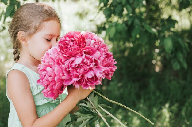 glückliches kleines Mädchen hält in ihren Händen einen Strauß rosa Pfingstrosenblüten in voller Blüte