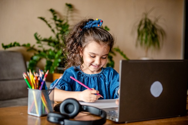 Glückliches kleines Mädchen in den Kopfhörern, die lächeln und Notizen im Notizblock machen, während sie mit Lehrer durch Video kommunizieren