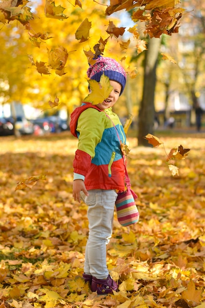 Glückliches kleines Mädchen in der hellen Kleidung, die mit Blättern in einem Stadtpark im Herbst spielt