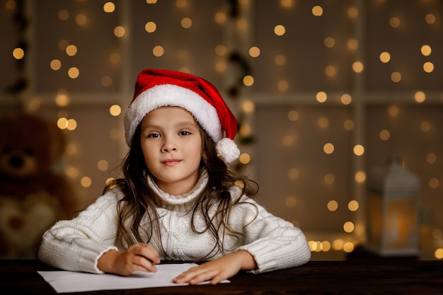 Glückliches kleines Mädchen in der Weihnachtsmütze, die Wunschliste oder Brief an den Weihnachtsmann schreibt