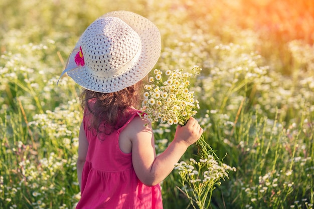 Glückliches kleines Mädchen in einem Strohhut mit einem Strauß Gänseblümchen Mädchen, das an einem sonnigen Sommertag auf einem Blumenfeld spazieren geht