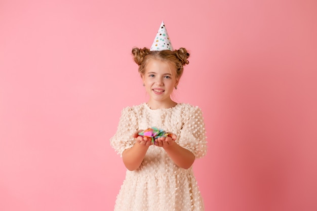 Glückliches kleines Mädchen in einer Geburtstagskappe bläst ihre Handflächen mehrfarbiges Konfetti auf einem rosa Hintergrund im Studio ab