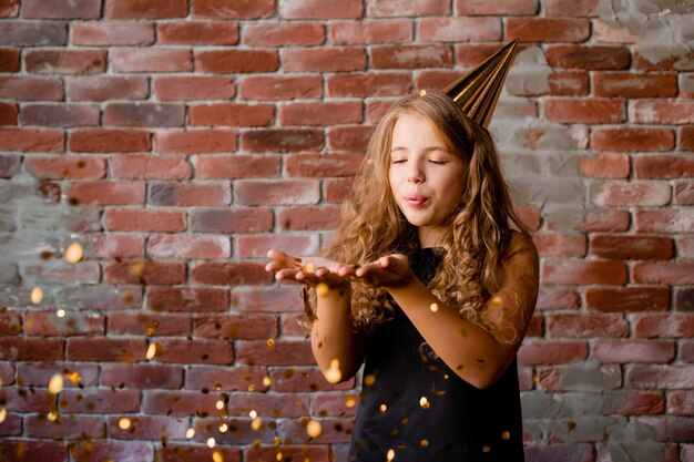 Glückliches kleines Mädchen in einer Kappe für ihren Geburtstag bläst weg von den Palmen des Konfettis