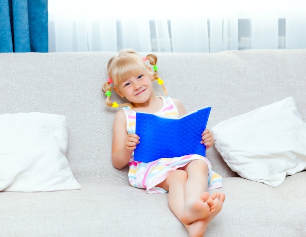 Glückliches kleines Mädchen mit blonden Haaren liest ein Buch auf einem Sofa im Wohnzimmer ihres Hauses. Fernunterricht.