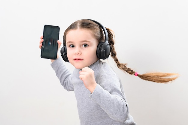 Glückliches kleines Mädchen mit drahtlosen Kopfhörern und Telefon Porträt eines Mädchens mit Zöpfen auf weißem Hintergrund mit Kopfhörern und Smartphone