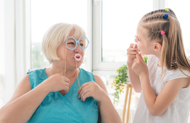 Glückliches kleines Mädchen mit Großmutter, die Brille und Lippen auf Stöcken im hellen Raum setzt