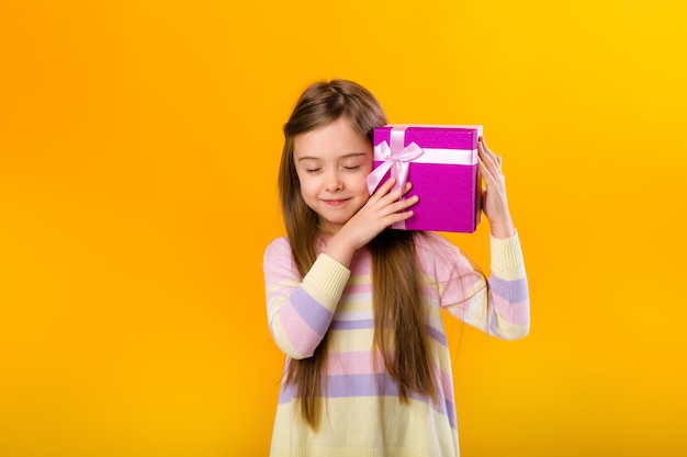 Glückliches kleines Mädchen mit langen Haaren, die eine rosa Geschenkbox auf einem gelben Raumisolat halten