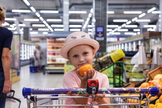 Glückliches kleines mädchen mit orange sitzt im einkaufswagen im obstladen, hat spaß und schaut in die kamera