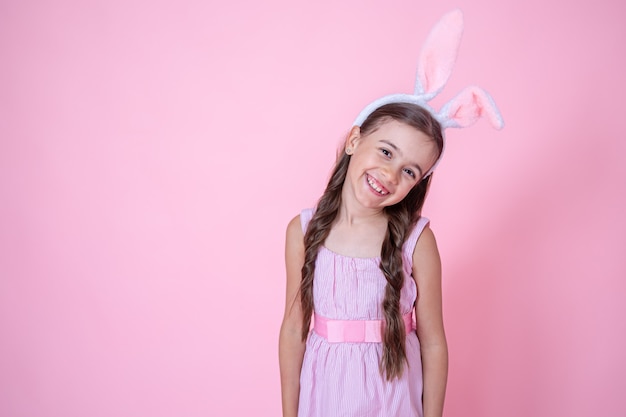 Glückliches kleines Mädchen mit Osterhasenohren, die auf einem rosa Hintergrundkopienraum des Studios aufwerfen. Osterferienkonzept.