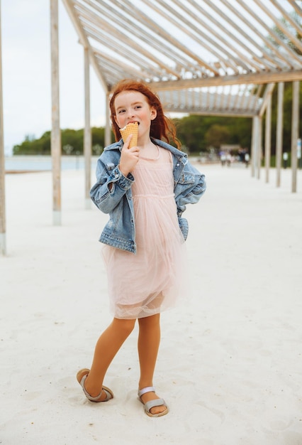 Glückliches kleines Mädchen mit roten Haaren isst in den Sommerferien Eis am Strand