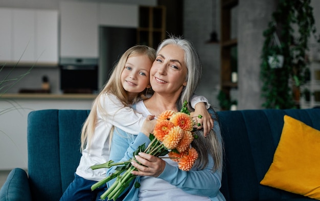 Glückliches kleines Mädchen umarmt kaukasische alte Großmutter gibt Blumen und Glückwünsche haben Spaß