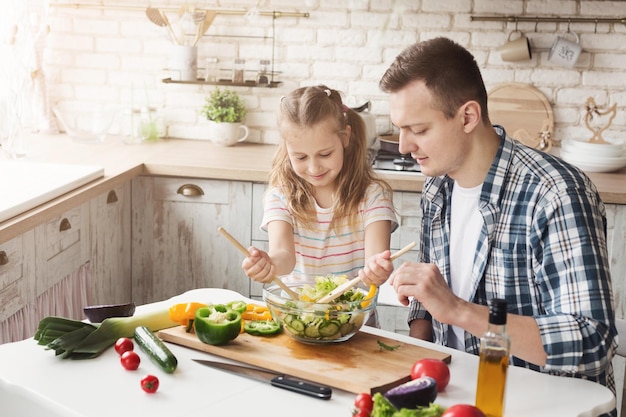 Glückliches kleines Mädchen und ihr Vater, die zu Hause kochen. Vater und Tochter machen Gemüsesalat und haben gemeinsam Spaß. Vatertag, Familie und gesundes Lebensmittelkonzept