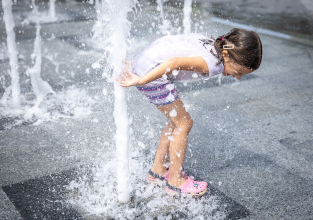 Glückliches kleines Mädchen unter dem Spritzwasser des Stadtbrunnens hat Spaß und entkommt der Hitze.