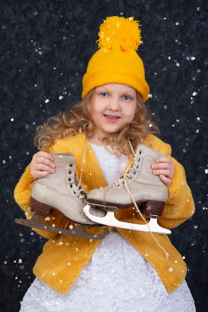 Glückliches kleines Mädchen unter dem weißen Schnee hält Schlittschuhe in den Händen und lächelt.