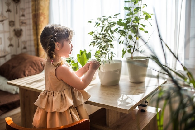 Glückliches kleines süßes Baby kümmert sich um Zimmerpflanzen. Mädchen, das Zimmerpflanzen zu Hause gießt und besprüht