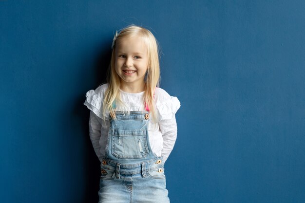 Glückliches Kleinkind hübsches Mädchen mit blondem Haar lächelnd, während auf blauem Wandhintergrund posierend