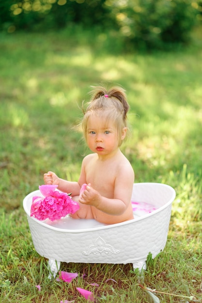 Foto glückliches kleinkindmädchen nimmt ein milchbad mit blütenblättern. kleines mädchen in einem milchbad. blumensträuße aus rosa pfingstrosen. baby baden. hygiene und pflege für kleine kinder.