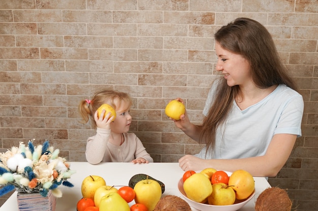 Glückliches Kleinkindmädchen und ihre schöne junge Mutter, die Frühstück macht und frische Früchte zusammen in einer Küche zu Hause isst