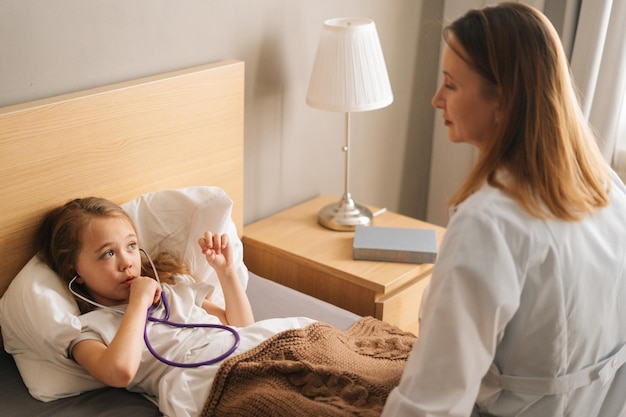 Glückliches krankes kleines mädchen, das mit stethoskop spielt, während es während des besuchs des kinderarztes in der medizinischen klinik im bett im schlafzimmer liegt